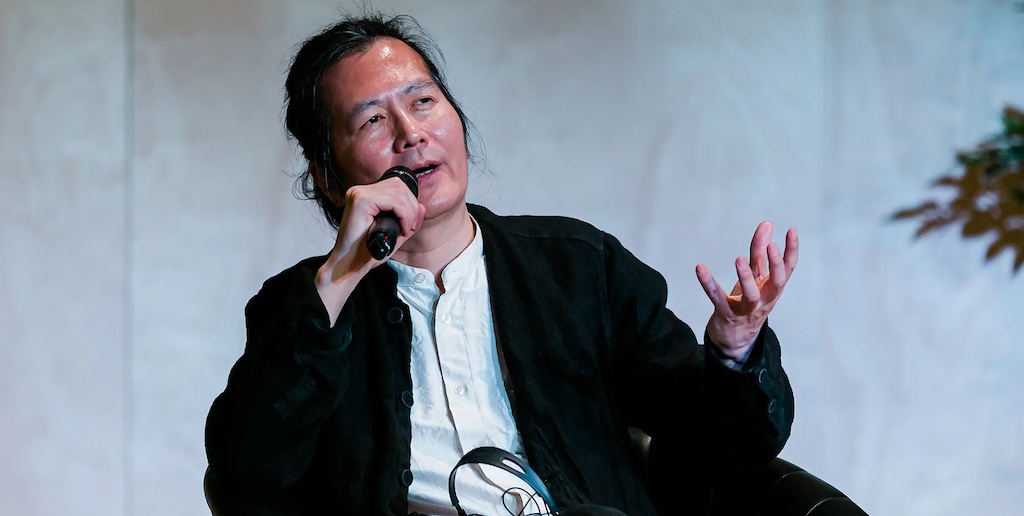 Byung-Chul dando una conferencia tiene el microfono cerca de su boca y gesticula con su mano izquierda