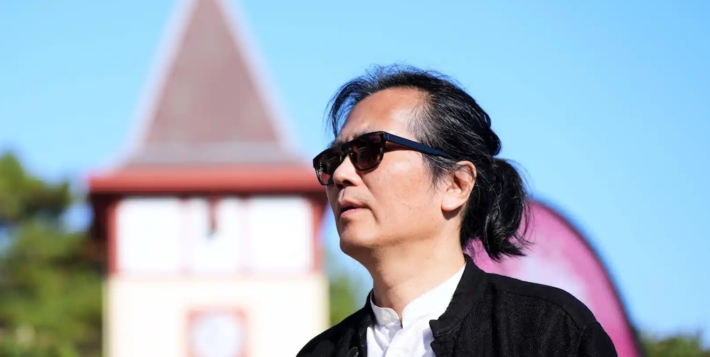 Fotografía del filósofo con lentes de sol en un día despejado, detrás de el se ve un edificación clásica
