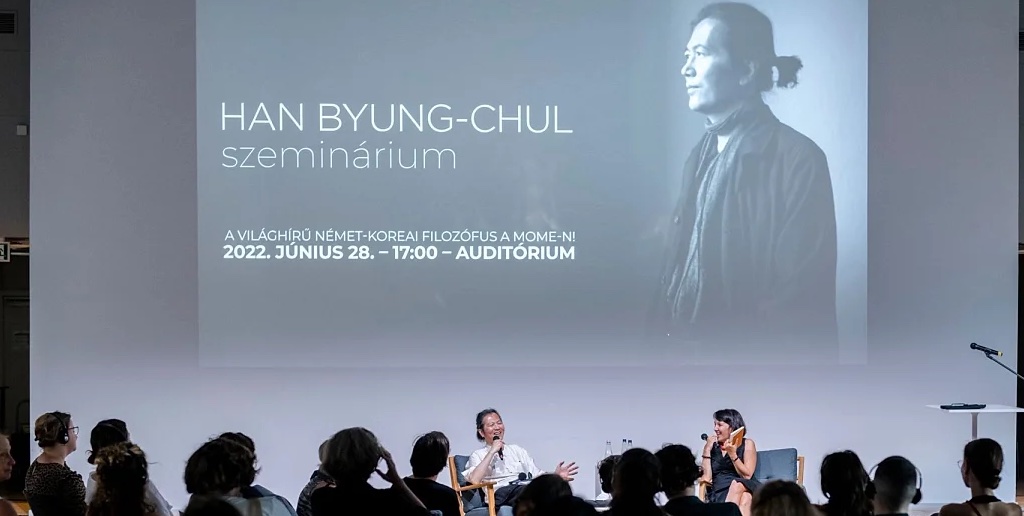 Byung-Chul Han sentado en un escenario junto a una presentadora dando una seminario, el filósofo se ríe junto con el público